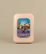 Image of travel tin with image of women in a bikini and hair towel reading a paper in a field of lavender and mint with cacti and mountains in the background