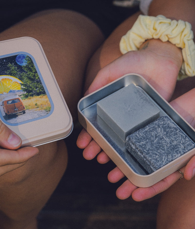 A set of hands holding an open travel tin showing black and grey shampoo and conditioner bars with a scrunchie on her wrist while another hand holds van life travel tin lid
