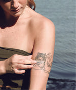Woman placing tuff stuff zinc sunscreen on a tattoo of a girl and a tiger with water in the background