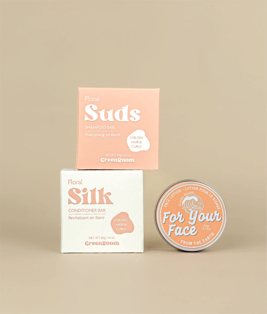 White with pink writing floral silk conditioner packing with the small pink with white writing floral suds shampoo bar packaging siting on top with a circular face lotion on its side to show orange label beside it