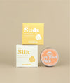 White with yellow writing sunny silk conditioner packing with the small yellow with white writing sunny suds shampoo bar packaging siting on top with a circular face lotion on its side to show orange label beside it