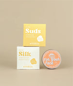 White with yellow writing sunny silk conditioner packing with the small yellow with white writing sunny suds shampoo bar packaging siting on top with a circular face lotion on its side to show orange label beside it