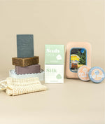 Fresh suds and silk boxes stacked with aloe mint soap stacked on oat milk latte soap and lavender zinc soap sitting on a white soap dish with a soap saver bag in front while on the other side the van life travel tin sits behind face lotion and zinc sunscreen in tins