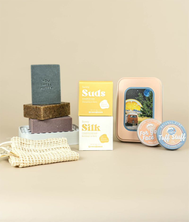 Sunny suds and silk boxes stacked with aloe mint soap stacked on oat milk latte soap and lavender zinc soap sitting on a white soap dish with a soap saver bag in front while on the other side the van life travel tin sits behind face lotion and zinc sunscreen in tins