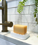 Dish soap block sitting on a white soap dish next to a brass tap with white subway tile and a plant in the background