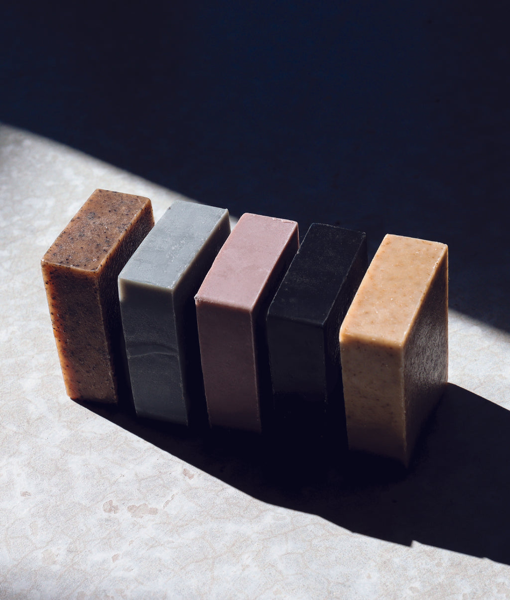 Oat milk latte soap, aloe mint soap, lavender zinc soap, charcoal and clay soap and seaweed and cedar soap lined up side by side in the sunshine with shadows around them