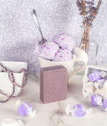 Lavender zinc soap with purple and white candies pouring out of a bowl, purple ice cream with sprinkles in a cup behind the soap with a spoon and purple and orange beads on the left hand side 