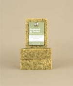 Two horizontally stacked seaweed and cedar soaps with a vertically placed seaweed and cedar soap on top with a green and white paper recycled tag held on by a metal push pin 