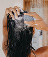 Woman holding small square fresh shampoo bar to scalp with suds in hair and shower pouring water in the back ground