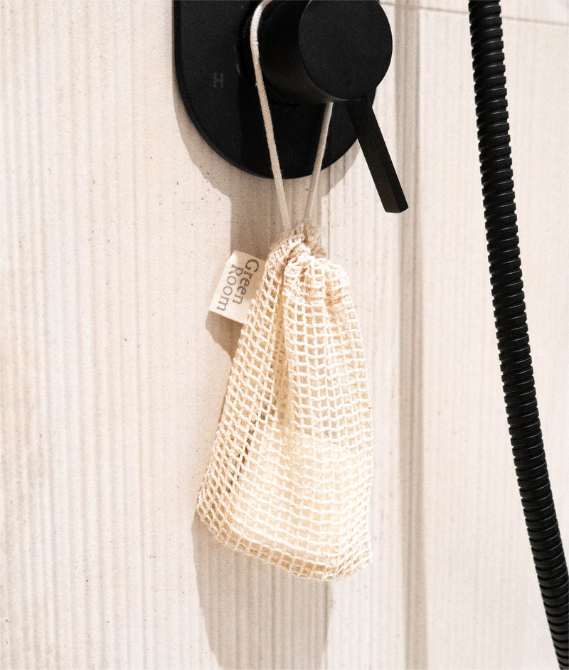 Natural mesh soap saver bag with green room tag hanging from black shower handle with shampoo bar in bag